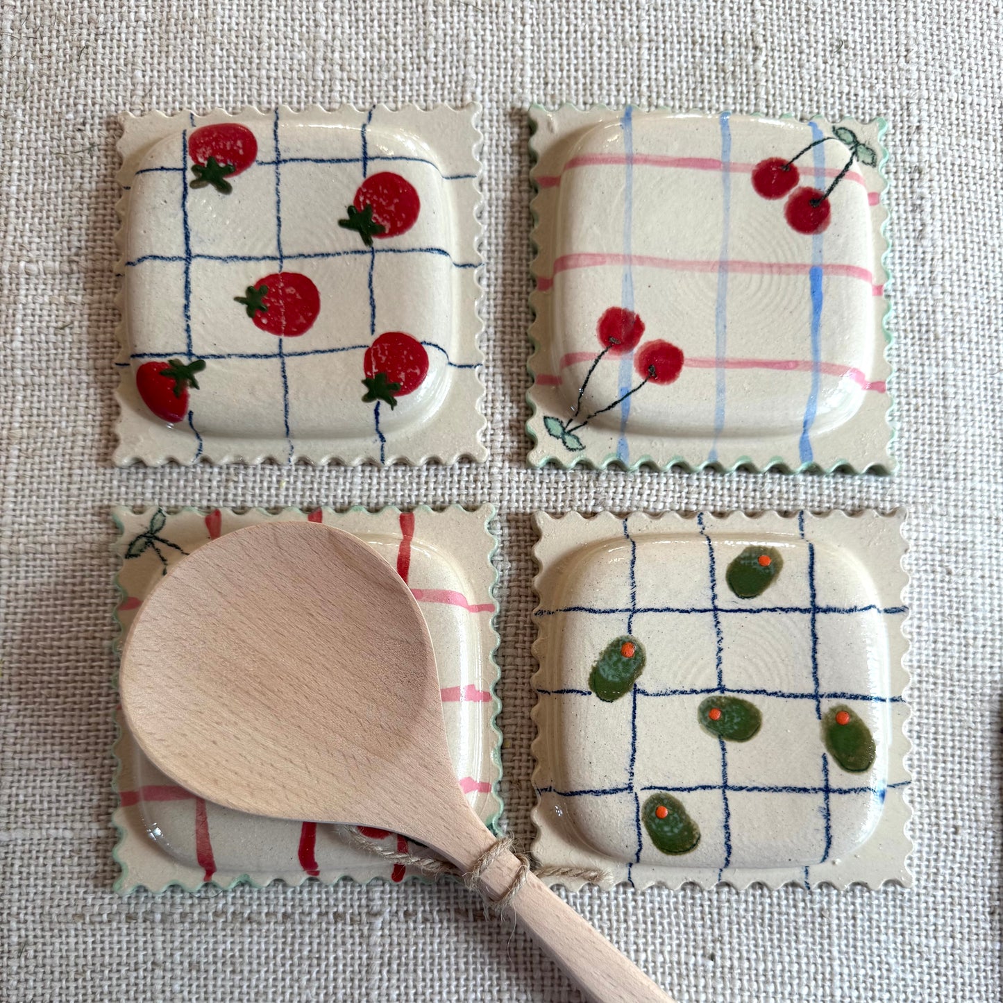 Four handmade ceramic ravioli spoon rests with cherry, tomato and olive designs on linen background