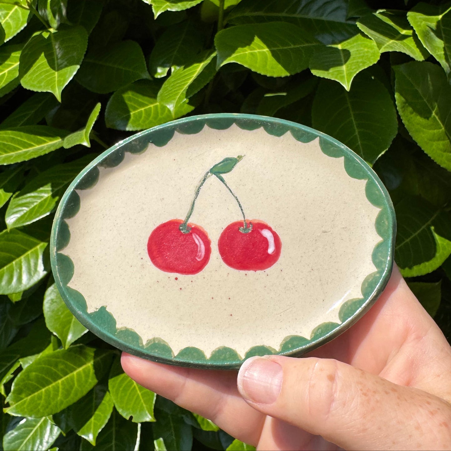 Handmade ceramic cherry design oval dish with sage green scalloped edge held outdoors